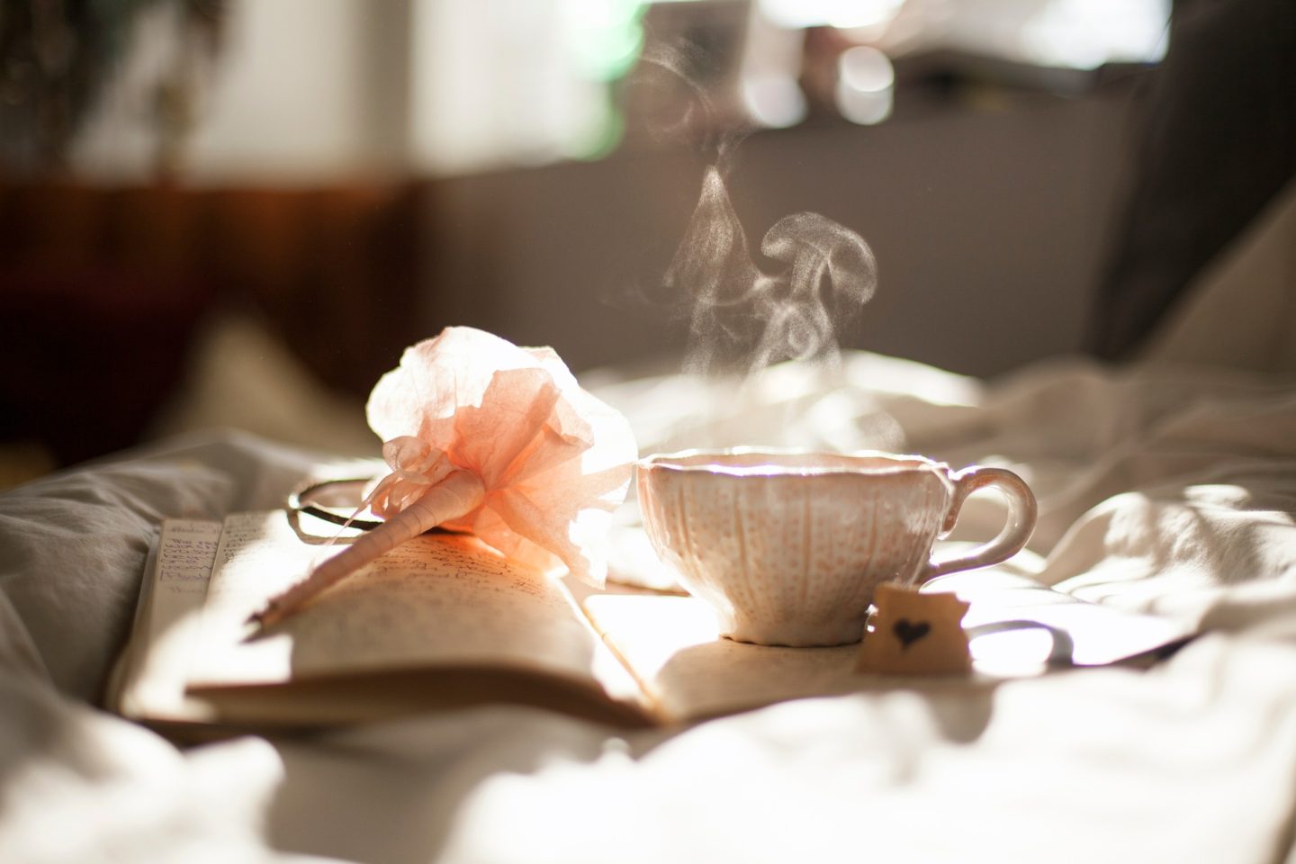 a hot cup of tea and a pen resting on top of a journal resting atop a duvet